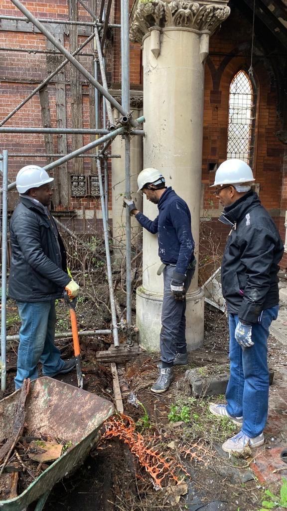 Three volunteers in hard hats clearing around a stone column